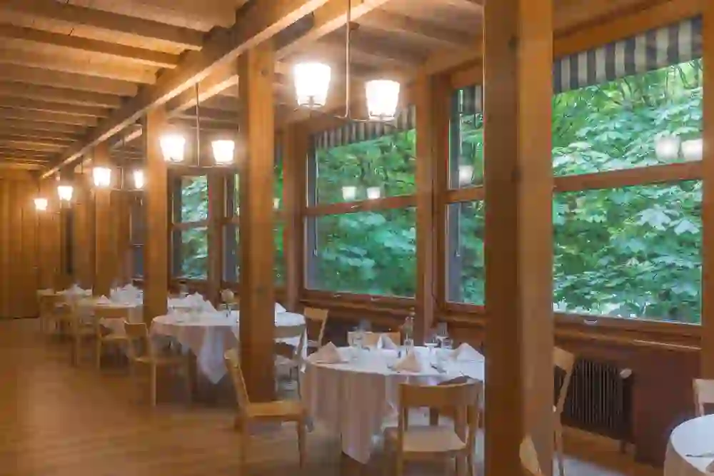 Una sala da pranzo luminosa con travi in legno e grandi finestre offre una vista sul verde lussureggiante all'esterno. Le tovaglie bianche e i tavoli apparecchiati con stile creano un'atmosfera tranquilla e invitante, circondata dalla natura.