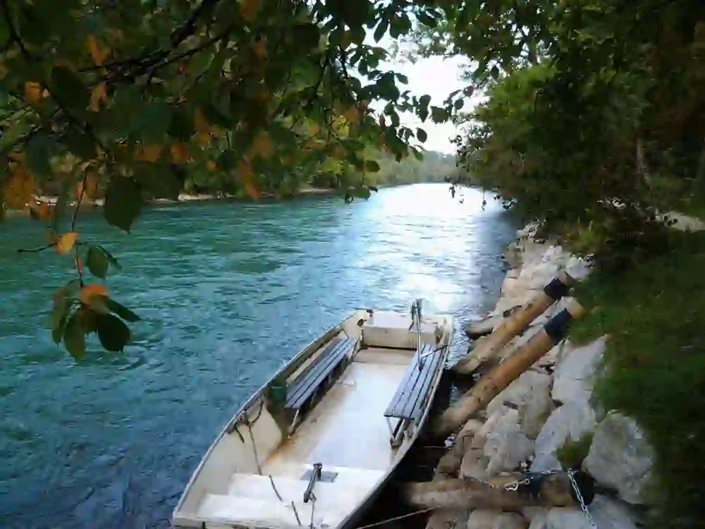 The photo shows a disused boat on the River Aare, embedded in a peaceful natural landscape. It is a calm day, the ripening leaves frame the scene and suggest a transition from summer to autumn. The boat belongs to the ferry pub in Elfenau on the Aare in Bern.