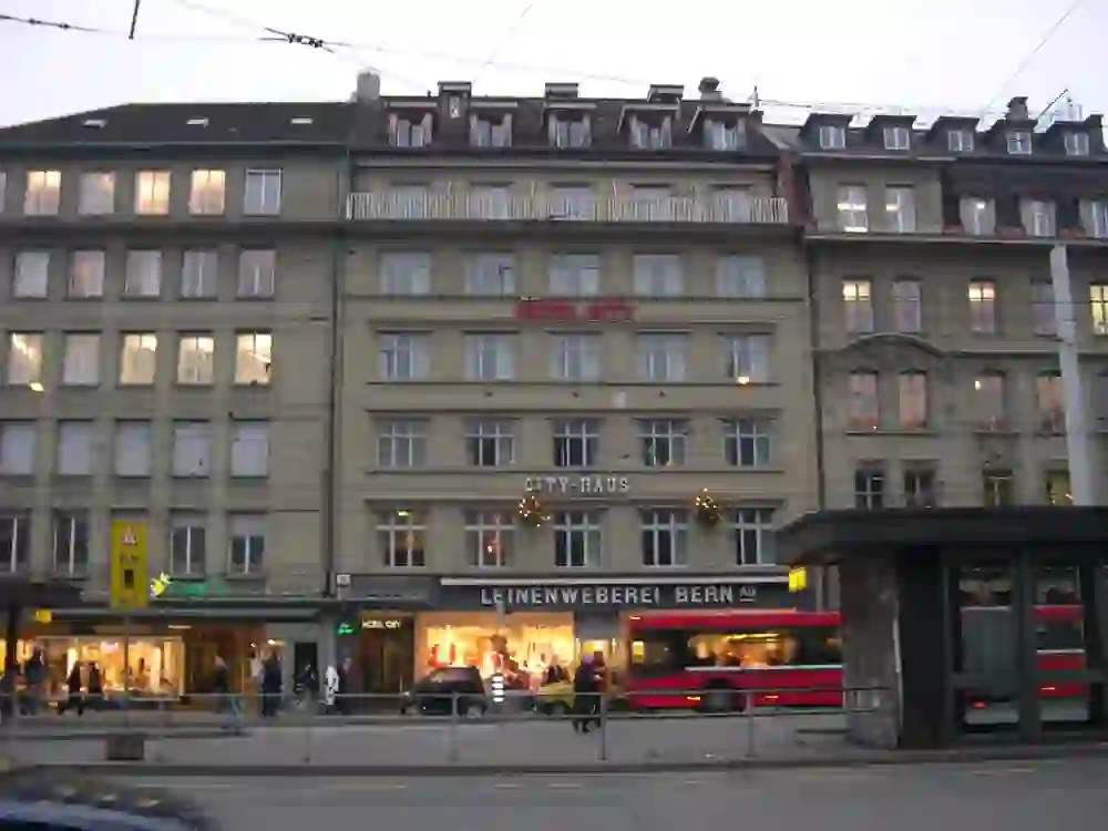 The image shows an urban street scenario with the Hotel City in the centre of the city of Bern The building has a classic façade with illuminated advertising for the hotel name. There are shops on the ground floor and passers-by on the pavement. A tram passes by, indicating a lively urban environment. The atmosphere seems mundane and dynamic, typical of a city centre in the evening or at dusk.