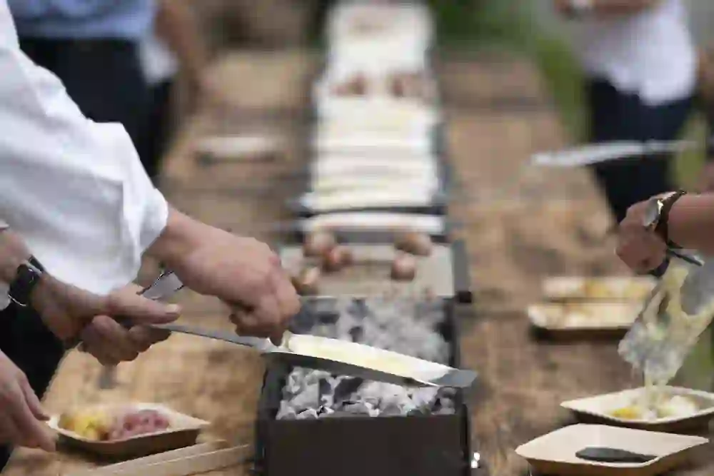 Des cuisiniers concentrés en train de préparer habilement la raclette sur un long gril, entourés de collègues conviviaux qui se réjouissent à l'idée de déguster la délicatesse chaude et fondante du fromage dans une atmosphère détendue et amicale.