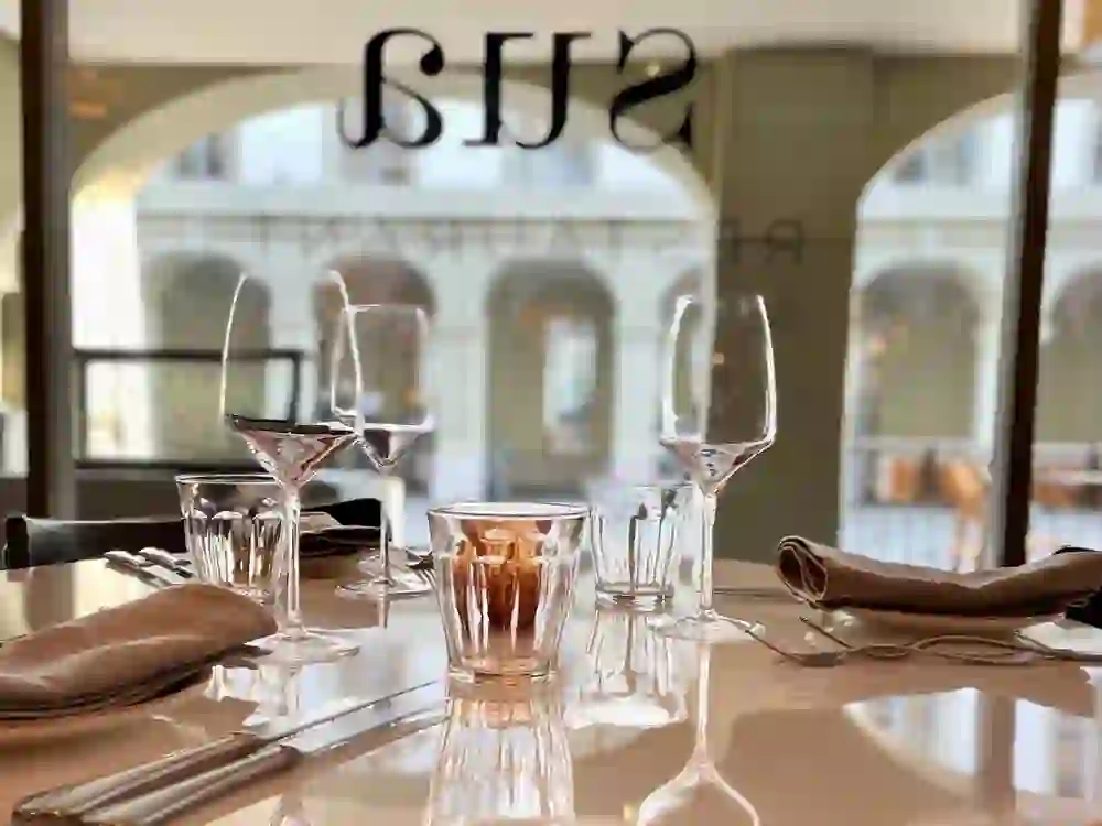 L'image montre une table élégamment dressée au restaurant Sua à Berne, à travers la fenêtre de laquelle on aperçoit un décor urbain, ce qui suscite un sentiment d'exclusivité et d'anticipation de l'expérience culinaire à venir.
