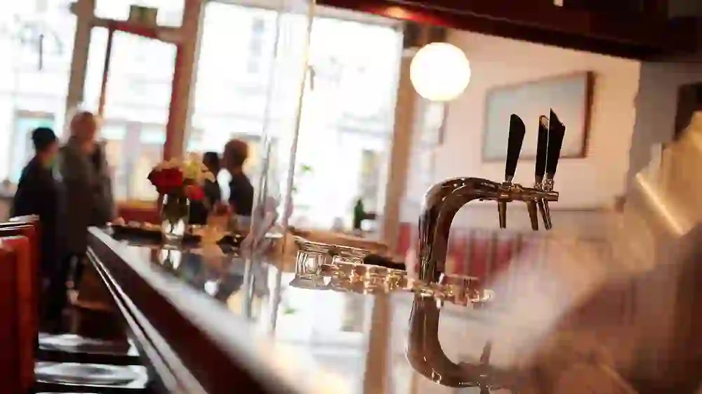 The picture shows a friendly restaurant scene with a focus on a shiny tap in the foreground. The reflection and natural light create a feeling of life and everyday flow. In the background, guests can be seen out of focus, contributing to the relaxed atmosphere.