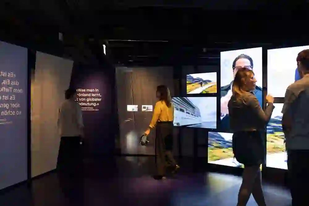 Das Bild zeigt eine immersive Ausstellung mit mehreren Bildschirmen, die Landschaften, ein Kreuzfahrtschiff und eine sprechende Person zeigen. Besucher bewegen sich durch den Raum, einige tragen Kopfhörer oder führen Gespräche. Beleuchtete Texttafeln enthalten Zitate, darunter: „Was man in Grönland lernt, ist von globaler Bedeutung.“ Die Szenerie wirkt modern und informativ, mit abgetrennten Bereichen und einer durchdachten Gestaltung. Die Ausstellung scheint sich mit Umwelt- oder Klimathemen zu befassen, möglicherweise im Kontext globaler Veränderungen.