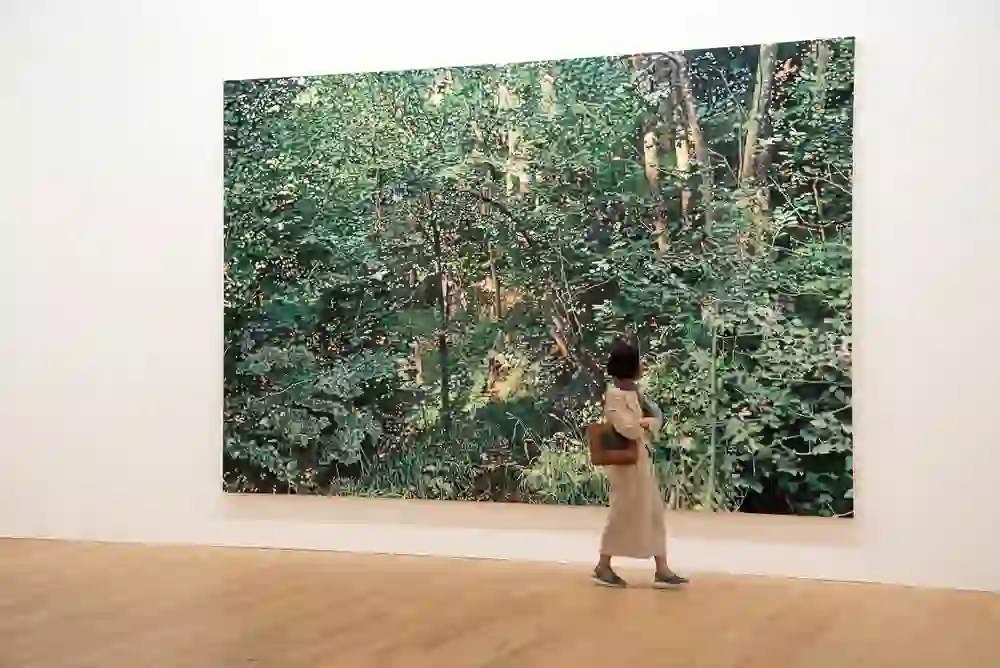 The picture captures a moment of quiet admiration in the Franz Gertsch Museum, in which a visitor stands in front of a life-size painting of a forest. The green colour tones of the artwork are reflected in the peaceful expression of the viewer, who seems to be in harmony with the nature of the painting.