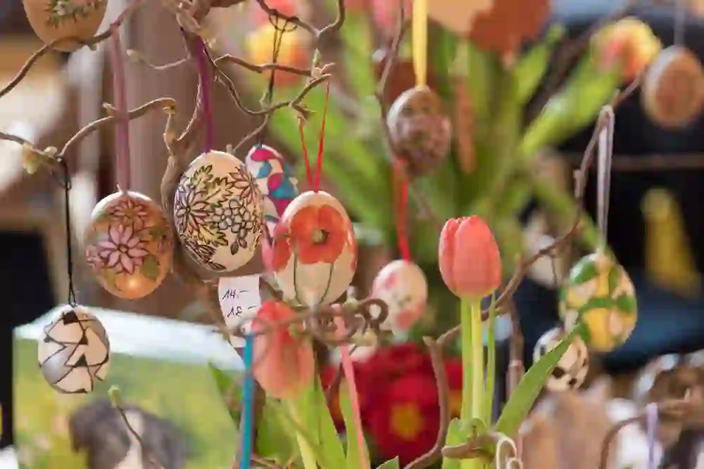 L'image montre un gros plan d'œufs de Pâques décorés et suspendus à des branches. Les œufs de Pâques sont peints avec art, certains avec des motifs floraux et d'autres avec des dessins abstraits. Au premier plan, on voit également des fleurs de tulipes, qui soulignent le thème printanier de l'image. En arrière-plan, on distingue d'autres plantes et décorations qui pourraient faire partie d'un marché festif ou d'une exposition. L'atmosphère semble colorée et joyeuse, en accord avec un marché de Pâques.