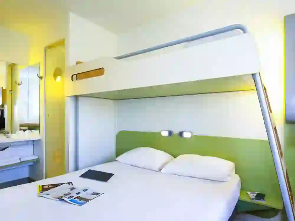 L'image montre une chambre d'hôtel à l'aménagement minimaliste, probablement d'un hôtel économique. L'ambiance semble pratique et efficace, avec un lit superposé qui permet de gagner de la place et une cabine de douche ouverte. L'aménagement est fonctionnel, ce qui donne une impression de sobriété et d'objectivité.