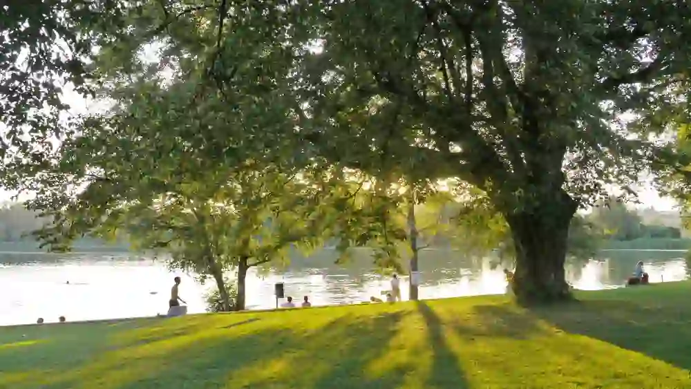 Das Bild zeigt einen friedlichen Abend am Burgäschisee in Aeschi. Menschen entspannen am Ufer, während die Sonne hinter den Bäumen untergeht und lange Schatten auf das leuchtend grüne Gras wirft. Ein Gefühl von Stille und Gelassenheit durchdringt diese sommerliche Szene am Wasser.
