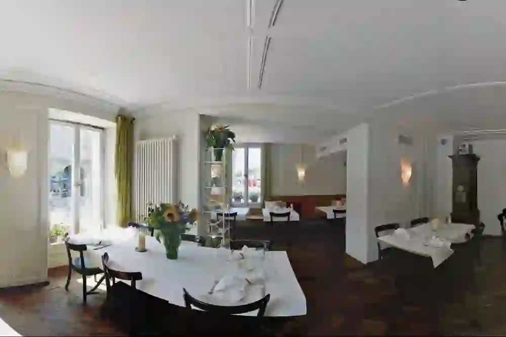 L'immagine panoramica mostra l'interno dell'elegante ristorante Ringgenberg di Berna. Ci sono diversi tavoli apparecchiati con tovaglie bianche. Ci sono luci decorative alle pareti e grandi finestre che lasciano entrare molta luce naturale. Il locale ha un aspetto classico con pareti bianche e soffitti in stucco. Una stufa in maiolica sul lato destro aggiunge un tocco tradizionale. Il pavimento in parquet aggiunge un'atmosfera elegante.