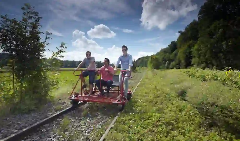Auf einer stillgelegten Bahnstrecke nahe Laupen radeln fröhliche Abenteurer auf einem Schienenvelo, umgeben von sommerlicher Natur und unter einem himmelblauen, wolkenverzierten Himmel.