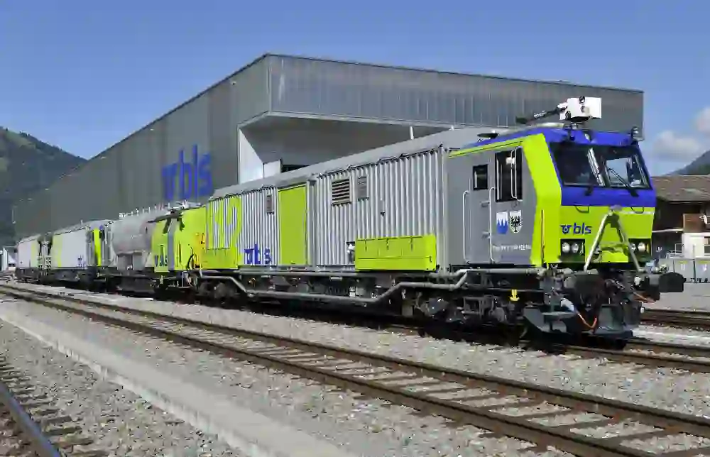 Un train de marchandises aux couleurs vives de la BLS est stationné sur des voies ensoleillées devant un bâtiment portant un grand logo BLS. La scène semble calme, propre et ordonnée, elle respire l'efficacité technique.