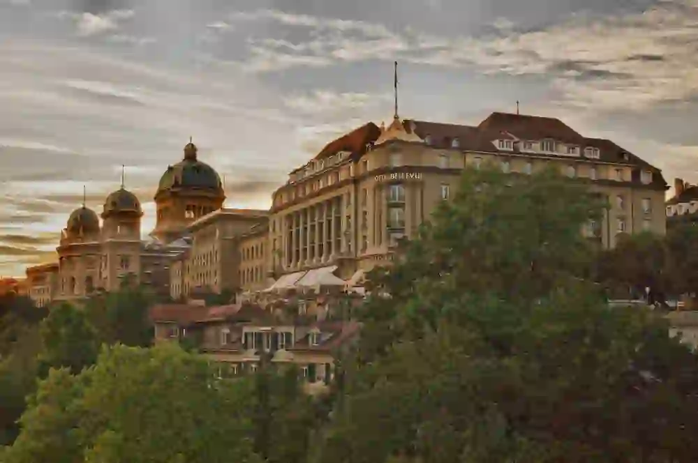 Das Bundeshaus und Hotel Bellevue in Bern bei Abenddämmerung, mit dramatischem Himmel und Bäumen im Vordergrund.