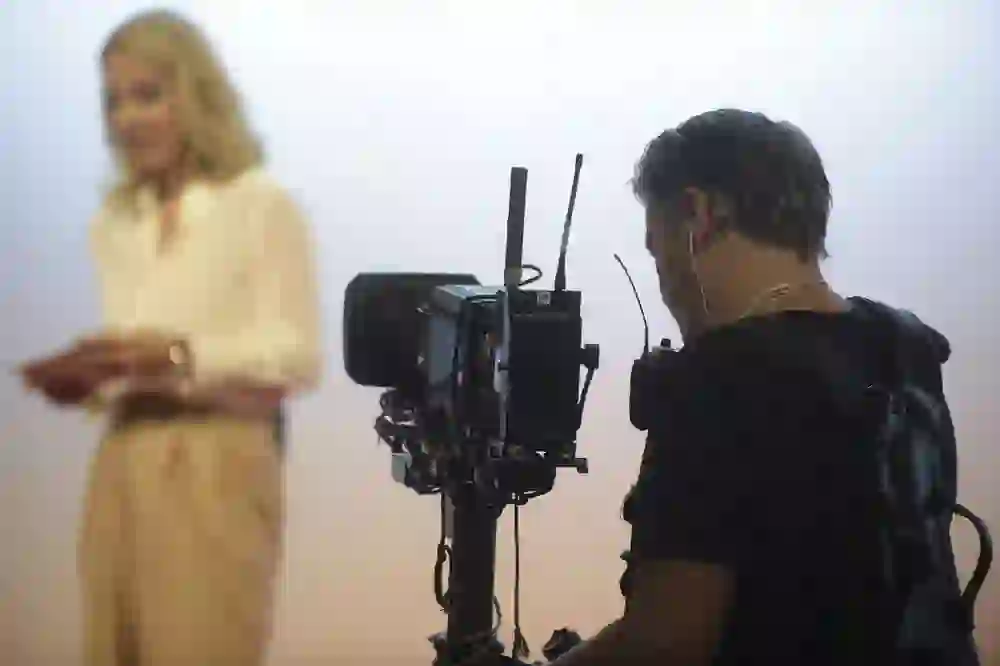 The picture shows an intimate film scene: a cameraman, concentrated and equipped with professional technology, films a woman reading a script in stylish clothing. The soft light creates a calm, focussed atmosphere filled with creative energy.
