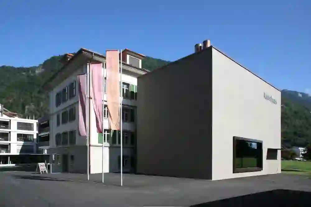The picture shows the Kunsthaus Interlaken on a bright day, surrounded by the tranquillity of the location and the majesty of the mountains. The clean lines and modern windows contrast with the traditional building next to it and convey a sense of contemporary elegance.