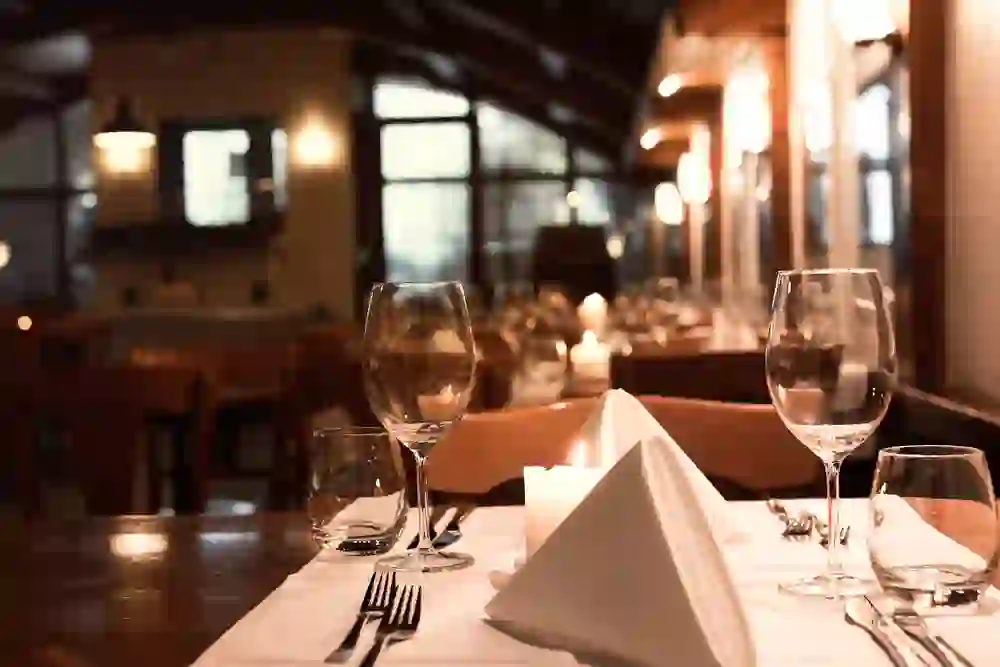 Das Bild zeigt einen gedeckten Tisch in einem Restaurant mit warmem Ambiente. Gläser und Besteck sind sorgfältig arrangiert, und Kerzenlicht trägt zur gemütlichen Stimmung bei. Der Fokus liegt auf dem Vordergrund, während der Rest des Restaurants sanft im Hintergrund verschwimmt, was eine intime und einladende Atmosphäre schafft.