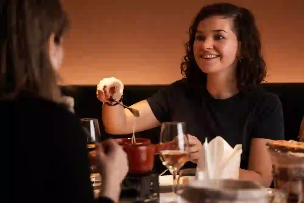 The picture shows a woman sitting at a table in a restaurant and eating fondue with a smile on her face. She appears to be having a pleasant time and enjoying the cosy atmosphere of the restaurant. The room is warmly lit, which together with the fondue creates an inviting and cosy atmosphere.