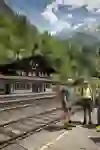 Quattro escursionisti si trovano in un'idilliaca stazione ferroviaria chiamata "Blausee-Mitholz", circondata da alberi rigogliosi e montagne imponenti. Sembrano eccitati e chiacchierano animatamente.