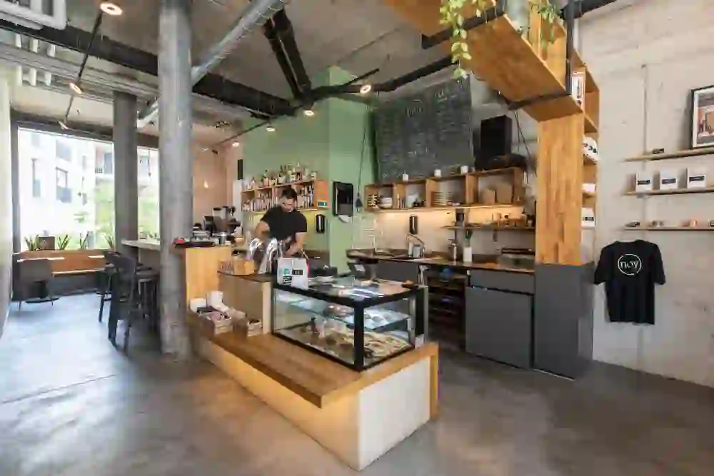 The image shows a modern café bar with an industrial aesthetic, emphasised by exposed concrete and exposed ventilation pipes. A barista concentrates on his work behind the counter, surrounded by a relaxed, welcoming atmosphere that invites you to linger.