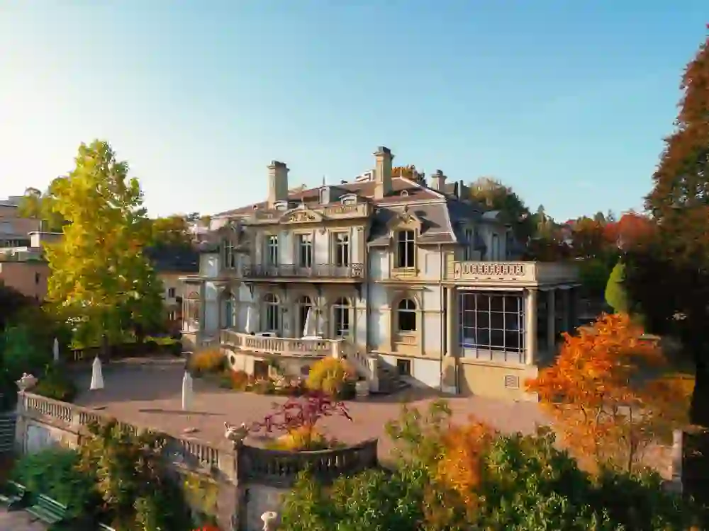 Das Bild zeigt eine prachtvolle, historische Villa umgeben von herbstlich gefärbten Bäumen. Das goldene Licht der tief stehenden Sonne taucht die Szene in eine warme, gemütliche Atmosphäre. Die farbenfrohen Blätter verstärken das Gefühl von Ruhe und Beschaulichkeit, als ob die Zeit für einen Moment stillsteht.