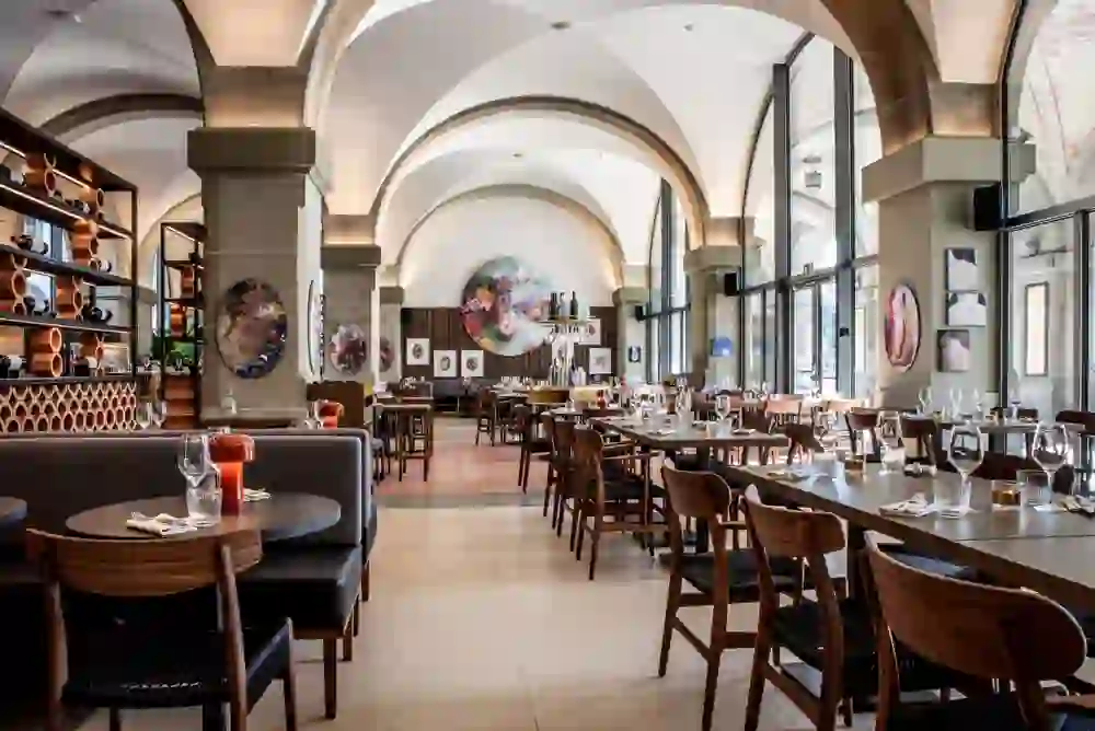 The photo shows the interior of a spacious restaurant with vaulted ceilings and large windows. The tables are finely laid and wine racks adorn the wall, creating an upscale and inviting atmosphere. Artwork on the walls adds a modern touch. It exudes calm and readiness for an elegant dining experience.