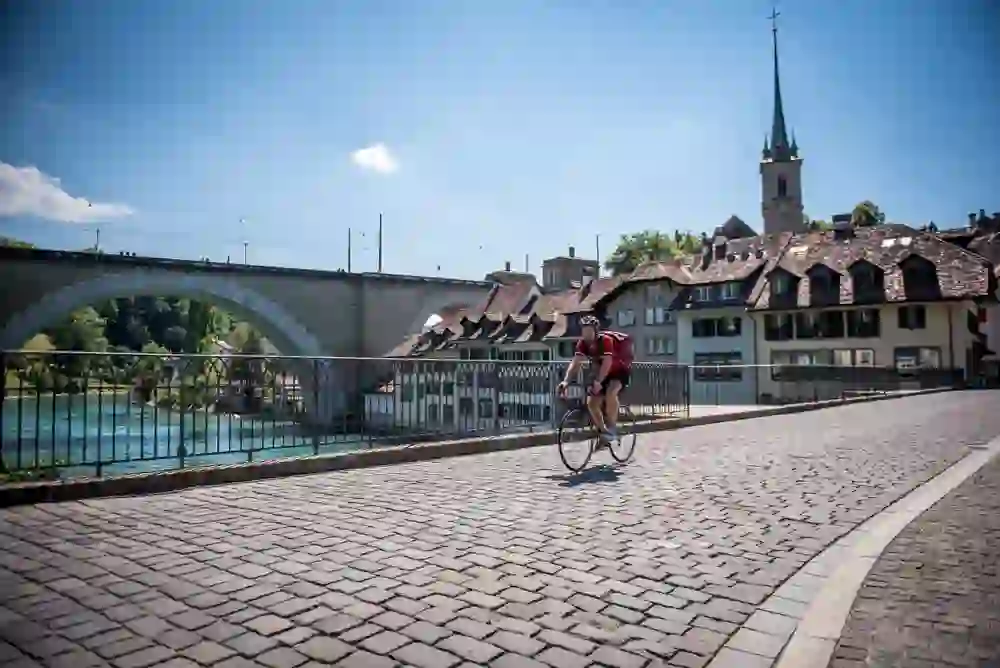 Ein Rennradfahrer überquert die gepflasterte Nydeggbrücke in Bern, im Hintergrund die historische Untertorbrücke und das türkisfarbene Wasser der Aare. Die Architektur und das klare Sommerwetter schaffen eine Kulisse, die sowohl das reiche Erbe der Stadt als auch die Lebendigkeit des modernen Lebens einfängt.
