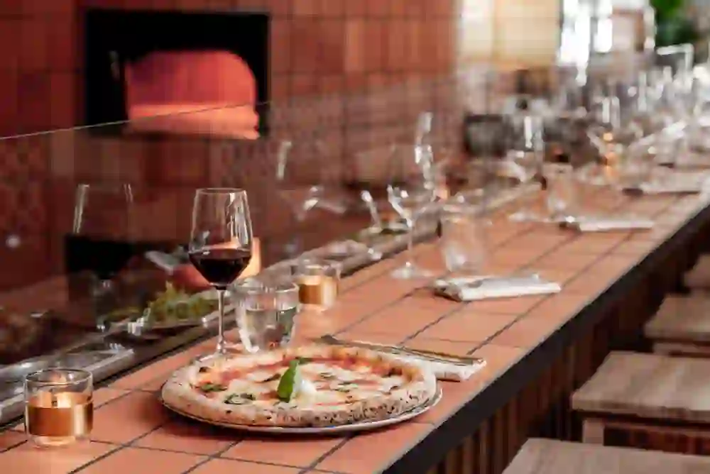 The picture shows a freshly prepared pizza on a restaurant counter with a backdrop of a glowing pizza oven. The table decorations, the red wine glass and the warm lighting create an inviting, cosy atmosphere, ideal for a relaxed culinary experience.