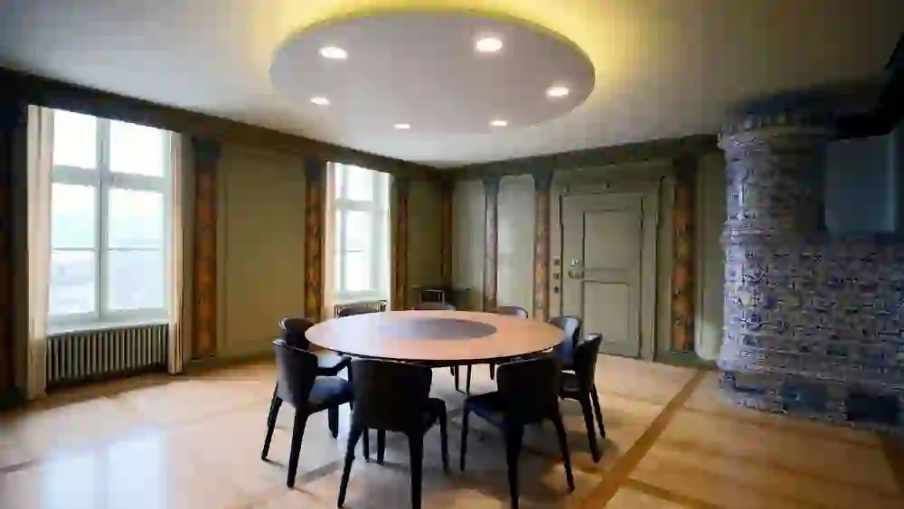 Das Bild zeigt einen eleganten Raum mit einer Kombination aus traditioneller und moderner Einrichtung. Im Zentrum steht ein runder Tisch mit schwarzen Stühlen. Über dem Tisch hängt eine große, moderne Deckenleuchte. An der Seite des Raumes befindet sich ein großer, aufwendig verzierter Keramikofen. Die Wände sind mit historischen Mustern verziert, und das große Fenster lässt viel Licht herein. Der Fußboden ist aus hellem Holz.