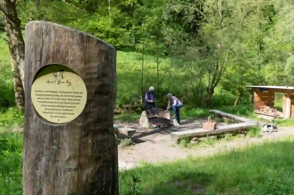The picture shows the idyllic barbecue site at the Mutzbach Falls in Riedtwil in a green, lively valley. A wooden plaque in the foreground presents a dedication or information, while in the background two people appear to be busy around a fireplace, perhaps preparing for a meal. A simple wooden shelter in the right-hand section of the picture offers protection and storage space for firewood.