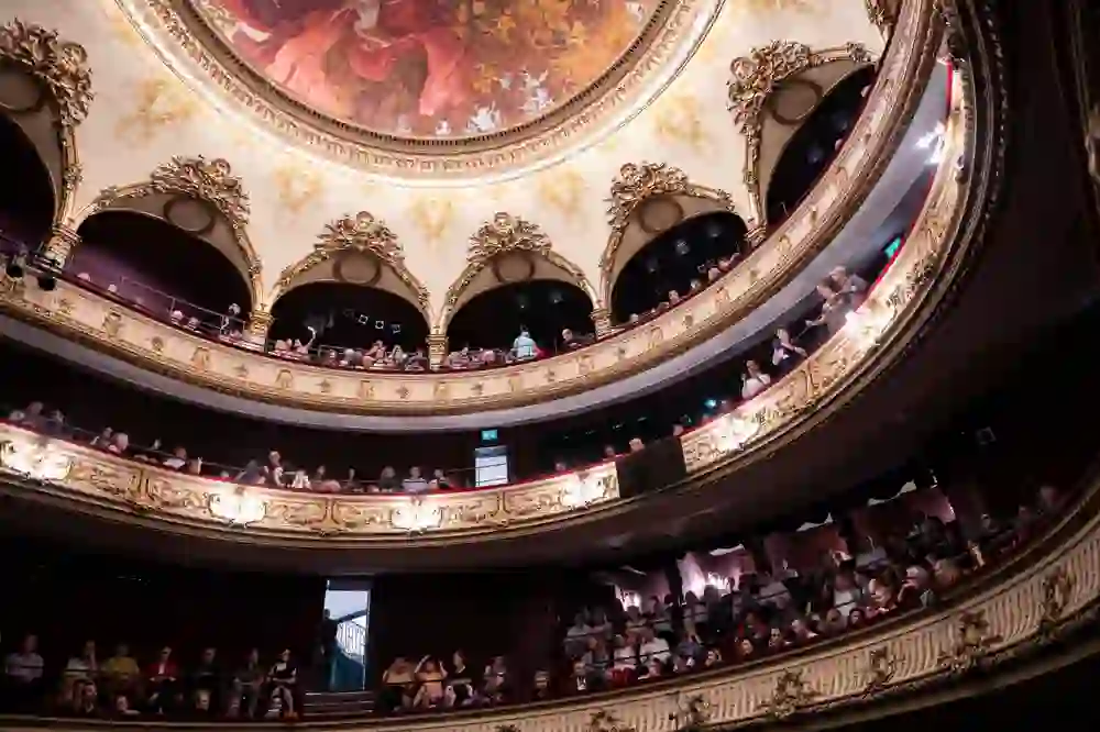 Das Bild zeigt einen prunkvollen Theatersaal mit aufwendig verzierten Balkonen und einer kunstvoll bemalten Decke. Die Zuschauer blicken gespannt auf das Geschehen. Die Atmosphäre ist feierlich und erwartungsvoll, während leise Gespräche die Vorfreude steigern.