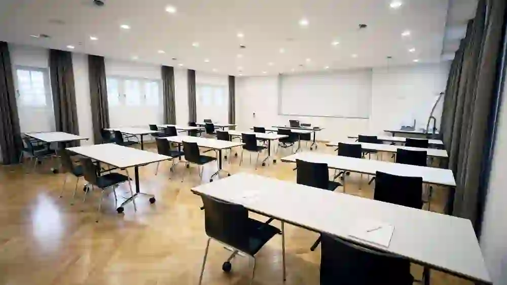 Das Bild zeigt einen hellen Seminarraum mit Holzparkett. Rechteckige Tische sind mit Stühlen um sie herum gruppiert und auf den Tischen liegt jeweils ein Blatt Papier und ein Stift. Die Wand im Hintergrund ist weiß und leer, womöglich für eine Projektion gedacht. Der Raum ist mit mehreren Deckenspots gut ausgeleuchtet und durch die Fenster fällt Tageslicht ein, was eine angenehme Lern- oder Arbeitsatmosphäre schafft.