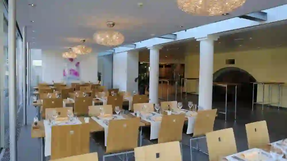 Auf dem Bild ist ein helles Restaurant mit einer modernen Einrichtung zu sehen. Mehrere Holztische mit aufgestellten Gläsern und Besteck sind für die Gäste vorbereitet. Über den Tischen hängen dekorative, kugelförmige Leuchten. Der Raum hat große Fenster, die für eine freundliche Atmosphäre sorgen, und einen dunklen Boden, der einen angenehmen Kontrast zum hellen Mobiliar bietet. Im Hintergrund ist ein Durchgang zu einem weiteren Raum erkennbar.