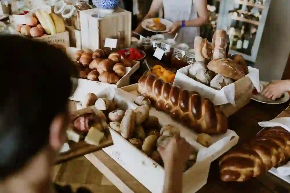 Auf dem Foto ist ein lebhaftes Brunch-Buffet mit einer Vielfalt an Backwaren zu sehen. Zu den gezeigten Speisen gehören verschiedene Brotarten, Croissants, und in der Mitte des Bildes fällt ein großes, geflochtenes Brot auf. Personen im Hintergrund bedienen sich am Buffet, was auf eine angenehme und gesellige Atmosphäre hindeutet.