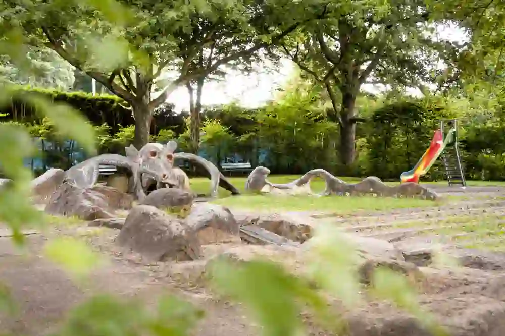 Das Bild zeigt einen Spiel- und Grillplatz in Weissenbühl bei Bern, der Kreativität und Abenteuer verspricht. Ein großer, skulpturaler Drache aus Beton windet sich durch die Szene, seine Formen und Strukturen bieten Kindern zahlreiche Möglichkeiten zum Klettern und Erkunden. Die lebendige Rutsche in der Form eines Drachenschwanzes fügt einen Farbakzent und ein Element des Spiels hinzu. Bäume spenden Schatten und bilden ein grünes Dach über diesem Ort des Lachens und der Freude.