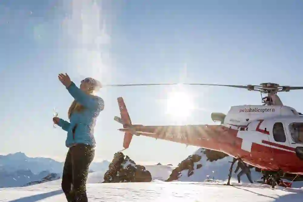 Eine Frau wirft lachend Schnee in die Luft, während sie ein Glas hält, mit einem roten Helikopter im Hintergrund, umgeben von einem schneebedeckten Bergpanorama und strahlendem Sonnenschein.