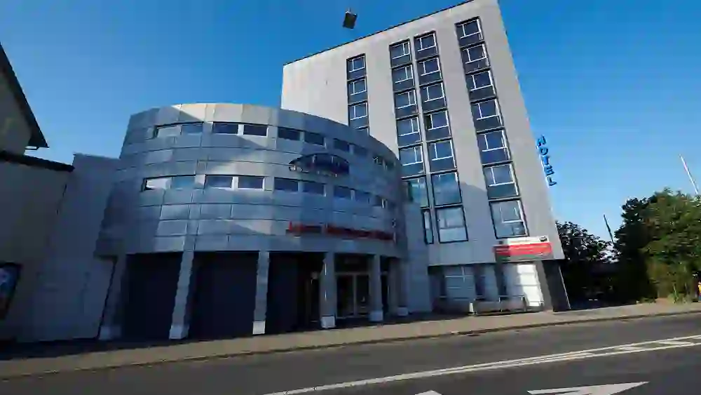 Das Foto präsentiert das moderne Gebäude des Hotel Ambassador in Bern, gekennzeichnet durch klare Linien und eine glatte Fassade, die eine urbane und zeitgenössische Atmosphäre unter einem strahlend blauen Himmel ausstrahlen.