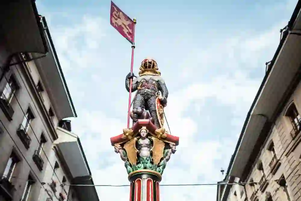 The photo shows a close-up of the Zähringerbrunnen in Bern with a bear in armour holding a flag against a background of city buildings and blue sky.