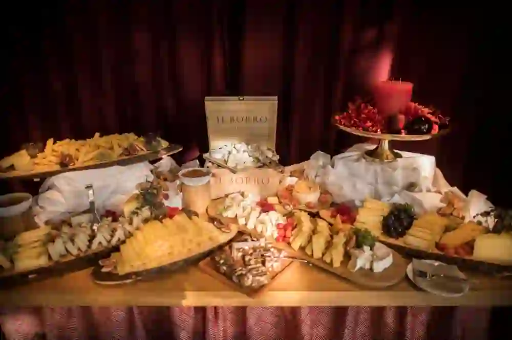 Auf dem Bild ist ein üppig angerichtetes Buffet mit einer Auswahl an Käsen, Trauben, Nüssen und anderen Snacks zu sehen. Es gibt verschiedene Käsesorten, die kunstvoll auf Holzbrettern präsentiert werden. Im Hintergrund ist eine weinrote Gardine, und auf einem Teller befindet sich eine dekorative rote Kerze umgeben von Blumen und Weihnachtsschmuck.