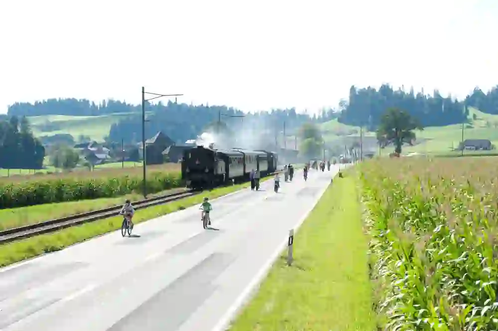 The picture shows an idyllic scene from the "Slow-Up" event in Emmental-Oberaargau: Cyclists of all ages enjoy a car-free road while a steam train leisurely passes them by. Rural serenity and a touch of nostalgia pervade the peaceful landscape.