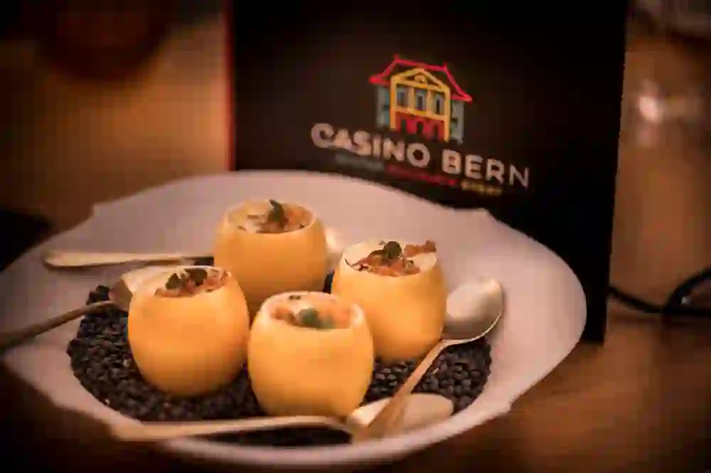 Das Bild zeigt vier runde, cremefarbene Desserts, die in Schalen angerichtet sind, welche aussehen wie ausgehöhlte Käsehälften. Sie sind auf schwarzen Linsen platziert, die als Dekoration auf einem weißen Teller dienen. Im Hintergrund ist ein Schild mit der Aufschrift "CASINO BERN" zu erkennen. Daneben liegt ein silberner Löffel.