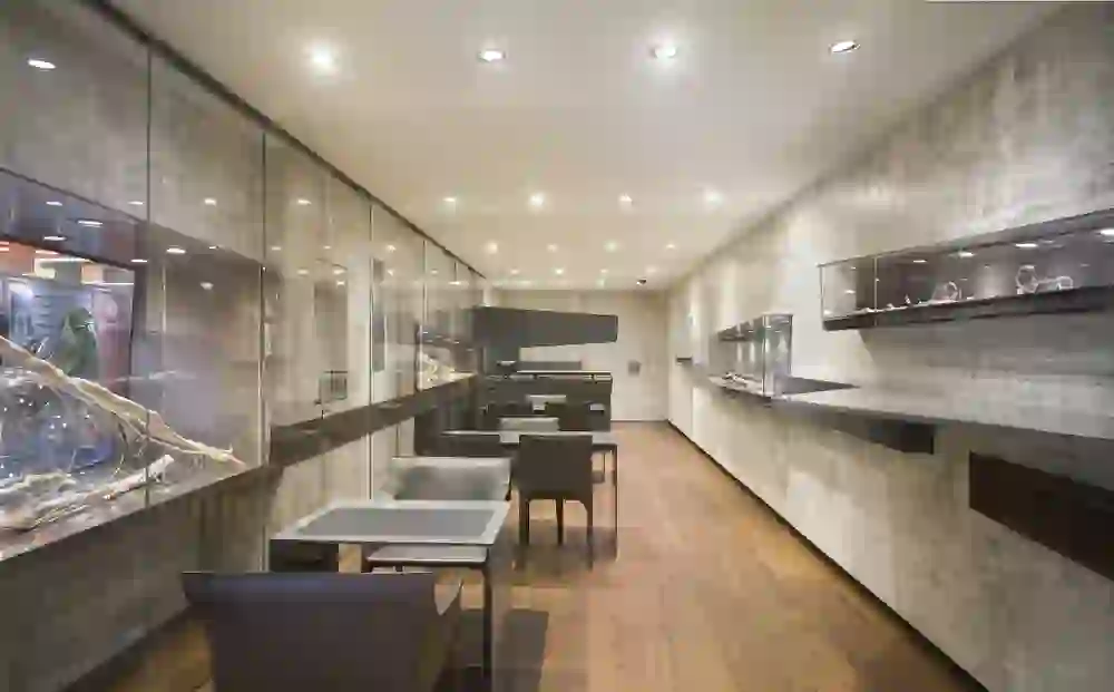 Das Bild zeigt das Innere eines eleganten Ladens, wahrscheinlich eines Juweliers, mit klaren Glasvitrinen an den Wänden, in denen Schmuckstücke ausgestellt sind. Der Raum hat eine moderne Gestaltung mit Beton- oder Steintapeten und ist mit Holzböden ausgestattet, was eine warme Atmosphäre schafft. Es gibt Tische und Stühle für Kunden.