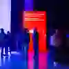 Les gens se rassemblent en groupes informels, discutent et semblent détendus. Des ambiances lumineuses colorées en bleu et en rouge plongent la scène dans une atmosphère moderne, tandis qu'un écran rouge invite à l'interaction.