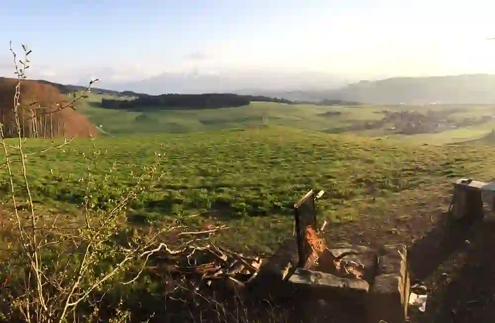 Auf dem Bild ist eine malerische Landschaft zu sehen, die von einer Anhöhe aus aufgenommen wurde. Im Vordergrund befinden sich ein kleiner Lagerfeuerplatz mit einem Gitter und einer Bank. Der Hintergrund zeigt weite, grüne Felder, einige Bäume und ein Dorf in der Ferne. Am Horizont sind Berge mit schneebedeckten Gipfeln sichtbar. Die Szene wirkt friedlich und idyllisch, beleuchtet von sanftem Sonnenlicht.