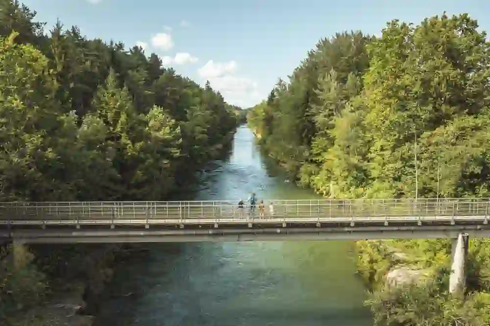 Das Bild zeigt eine Brücke über einen Fluss, der von dicht bewaldeten Ufern gesäumt ist. Auf der Brücke stehen vier Personen, die in den Fluss blicken. Die Vegetation ist üppig und grün, und der Himmel ist klar mit ein paar kleinen Wolken. Die Szene vermittelt eine ruhige und idyllische Atmosphäre in einer ländlichen Gegend.