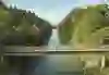 L'immagine mostra un ponte su un fiume costeggiato da sponde densamente boscose. Quattro persone sono in piedi sul ponte e guardano il fiume. La vegetazione è verde e rigogliosa e il cielo è limpido con qualche piccola nuvola. La scena trasmette un'atmosfera tranquilla e idilliaca in una zona rurale.