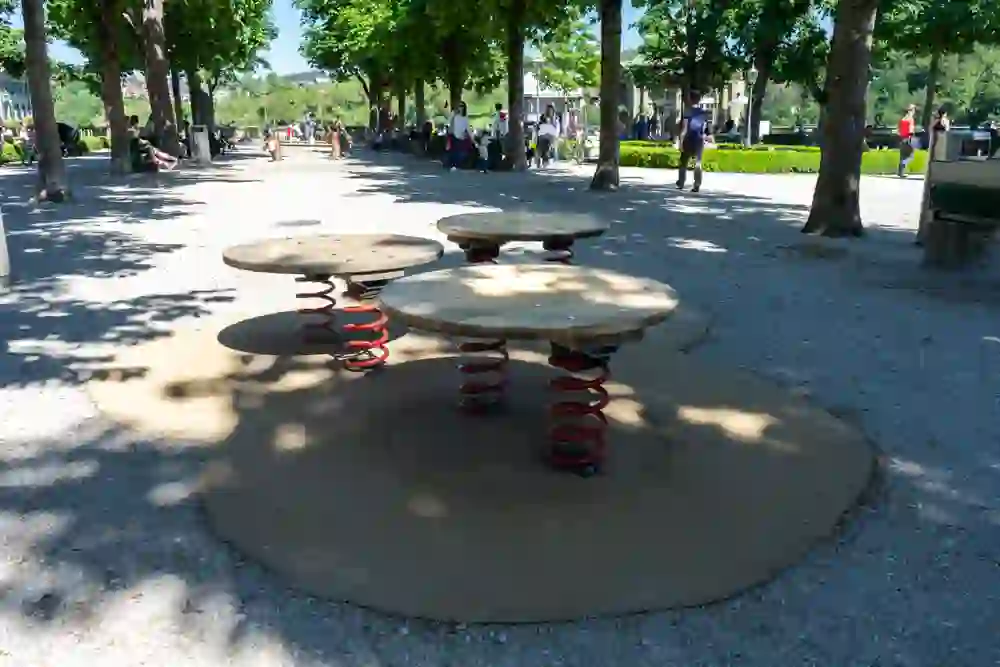 Das Bild zeigt einen lebendigen Spielplatz unter sommerlichem Sonnenschein, betont durch die Schatten der Bäume. Runde, federnde Spielgeräte laden zum dynamischen Spielen ein, während im Hintergrund Menschen entspannt flanieren, was eine unbeschwerte Stimmung erzeugt.