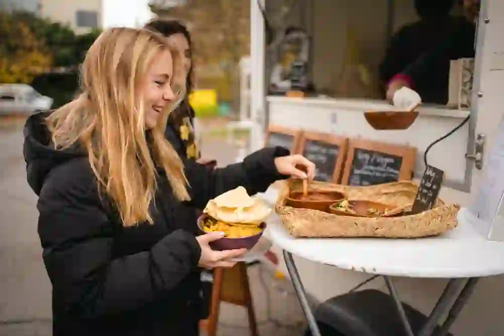 Eine junge Frau lächelt, während sie an einem Take-Away-Stand eine Schale mit köstlichem Curry füllt. Die warme und einladende Atmosphäre vermittelt Freude und Genuss, während sie das frisch zubereitete Essen serviert bekommt.