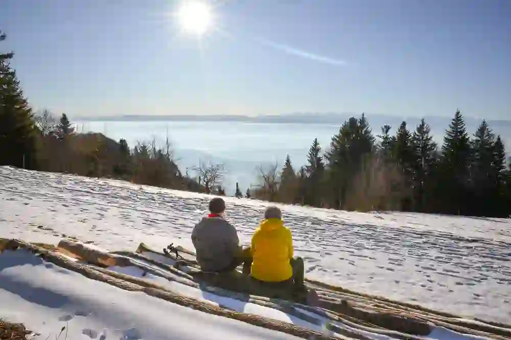 Zwei Personen sitzen friedvoll auf einem Baumstamm in einer verschneiten Landschaft, genießen die Wintersonne und blicken auf eine nebelverhangene Ferne mit Bergsilhouetten. Es strahlt Ruhe und Verbundenheit mit der Natur aus.