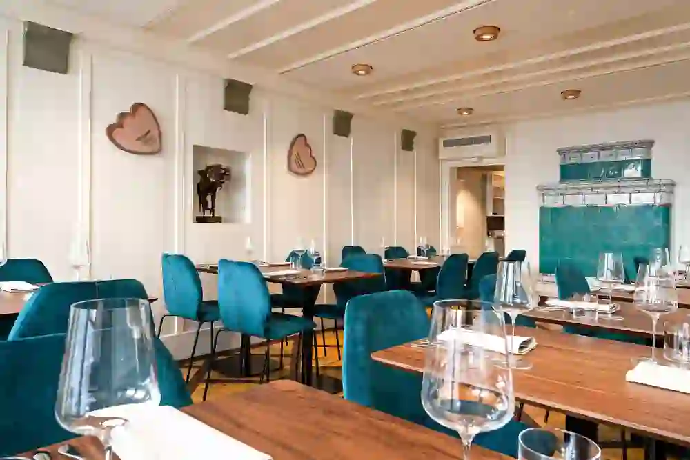 Das Foto präsentiert das helle und modern eingerichtetes Restaurant Zoe in Bern. Die lebendige Farbe der türkisblauen Samtstühle bildet einen schönen Kontrast zu den warmen Holztischen und der weissen Wandgestaltung. Die kunstvoll gestaltete Kachelofen in Grün setzt einen traditionellen Akzent in diesem ansonsten zeitgenössisch gestylten Raum. Die stilvolle Beleuchtung und das durchdachte Design schaffen eine ansprechende Atmosphäre, die zum Verweilen und Geniessen einlädt.