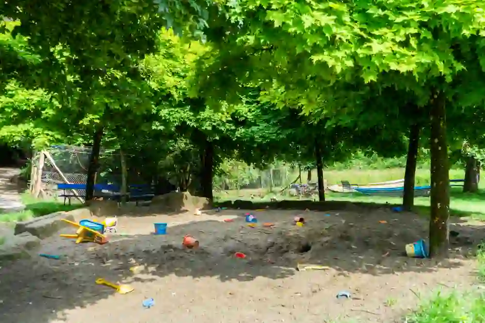 Das Bild zeigt einen ruhigen, sonnenbeschienenen Sandspielplatz unter dem Schatten grüner Laubbäume, verstreut mit bunten Spielzeugen. Die Atmosphäre ist friedlich und einladend, perfekt für ein entspanntes Spielen in der Natur.