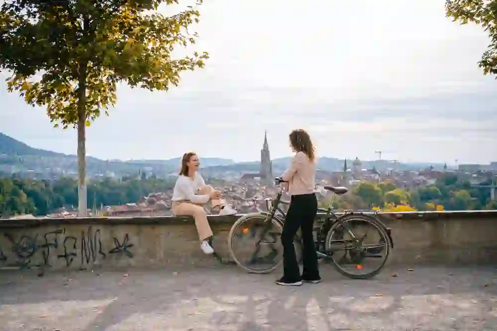 Two women are chatting in the rose garden in Bern, one sitting on the wall, the other standing next to it on her bike. They are enjoying the view of the city with Bern Cathedral in the background, embedded in the peaceful atmosphere of a spring day. The scene is characterised by serenity and the beauty of the moment.
