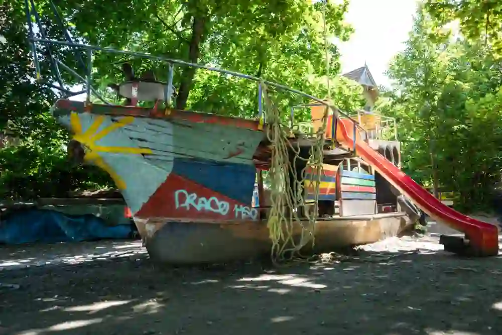 Das Bild zeigt ein farbenfrohes Spielplatzschiff umgeben von grünen Bäumen. Das Schiff trägt kreative Malereien und bietet mit einer Rutsche und Seilen ein lebhaftes Abenteuergefühl für Kinder. Ein Ort voller Fantasie und Spaß.